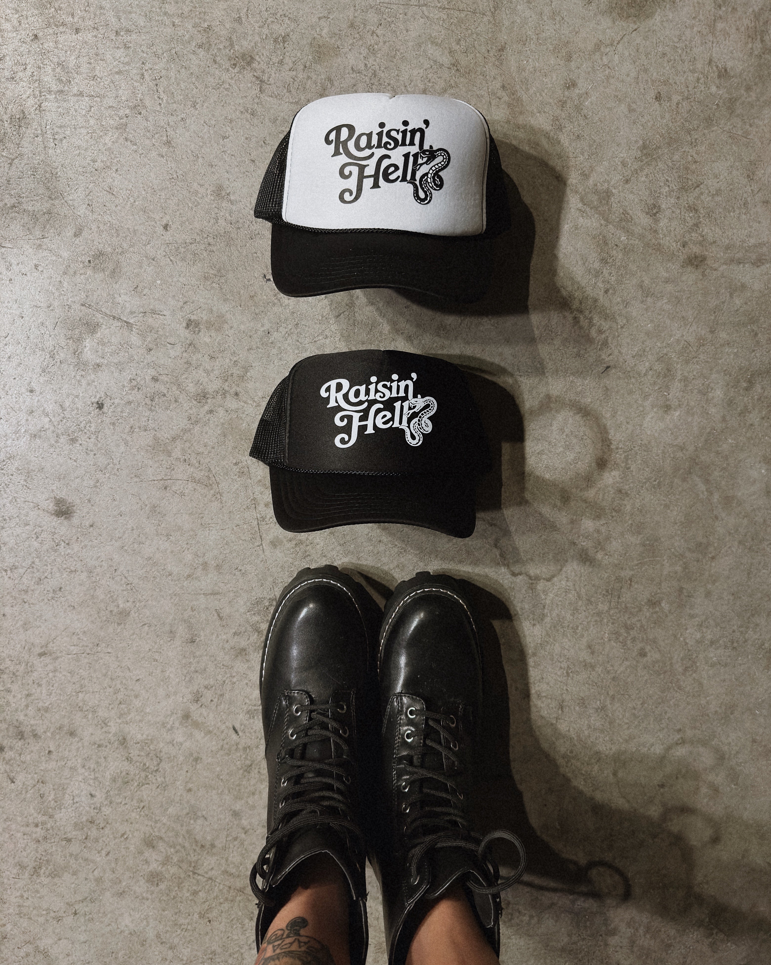 Two trucker hats, one black and one white, with the words “Raisin’ Hell” on a concrete ground with black combat boots