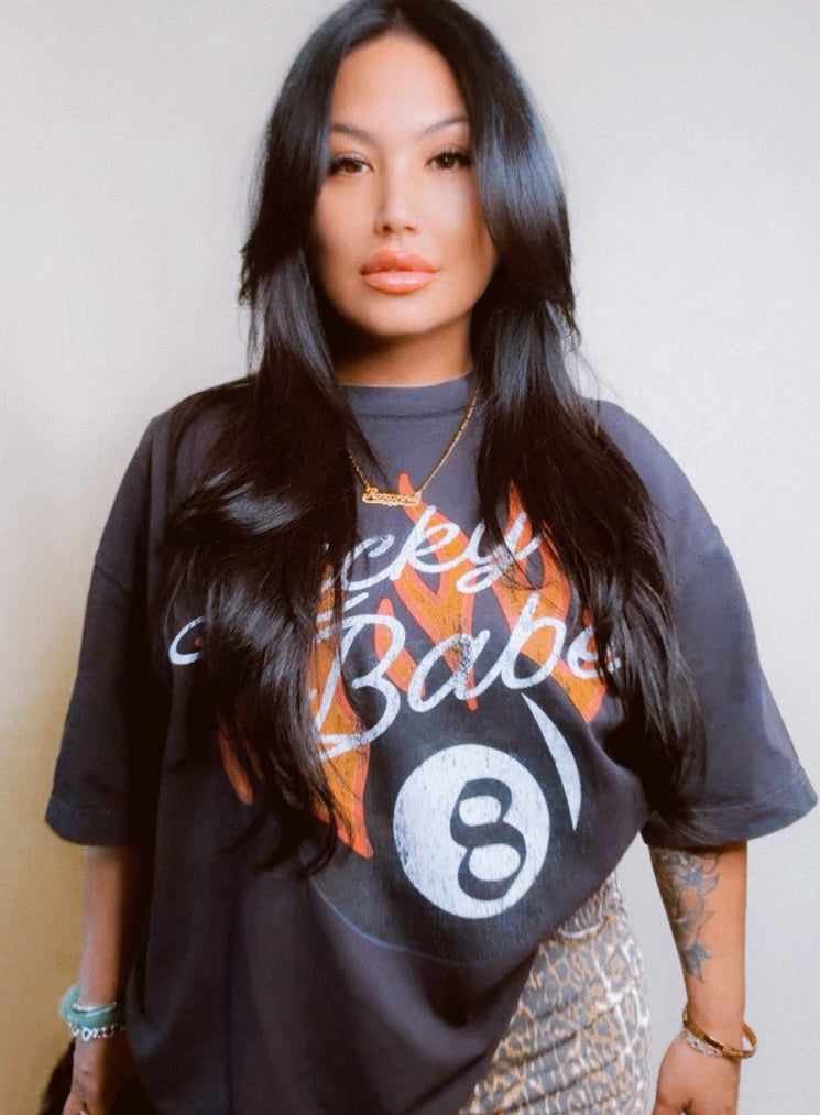 Women wearing a tee with a bold graphic of an 8 ball on fire with the words “Lucky Babe” in a script font.